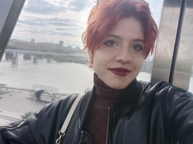 LadyIlona with red hair, dark lipstick, nose ring, and turtleneck, wearing black leather jacket, smiling, standing by window with city and river in background. Sheer selfie.