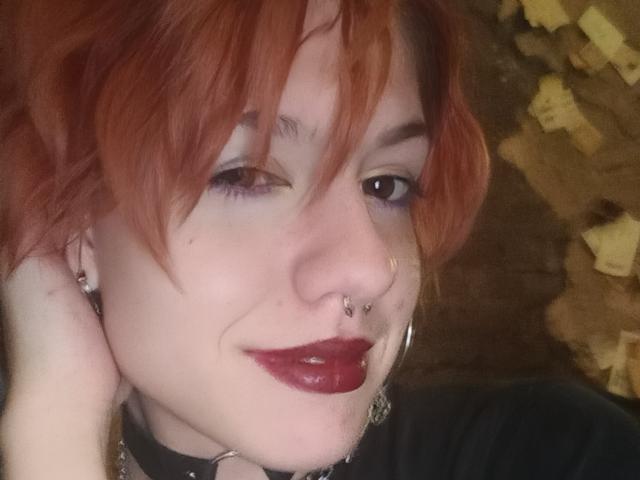 LadyIlona with red hair, nose piercings, and dark red lipstick, wearing a black choker and earrings, smiling slightly. Her hand is near her head.