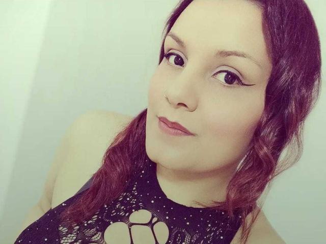ScarlettMancini with red braided hair, black lace top with cutout, eyeliner, nose ring, and subtle pink lipstick, looking slightly to the side, smiling gently.