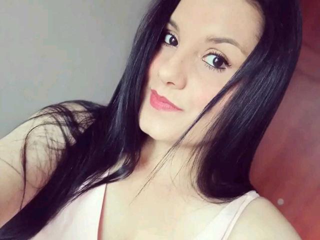 ScarlettMancini has long black hair, light skin, and pink lips, wearing a pink top, with small tits, looking at the camera with a slight smile.