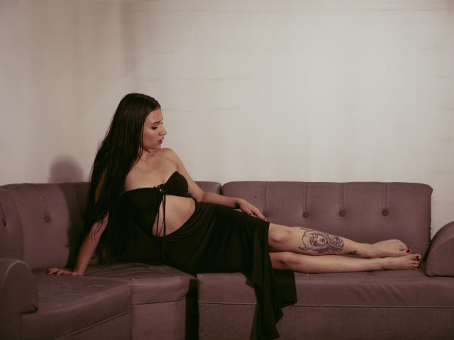 LizzeJoy with long black hair, lying on a purple couch in a black dress with side cutouts, showing a thigh tattoo, and resting her arm, looking to the side.