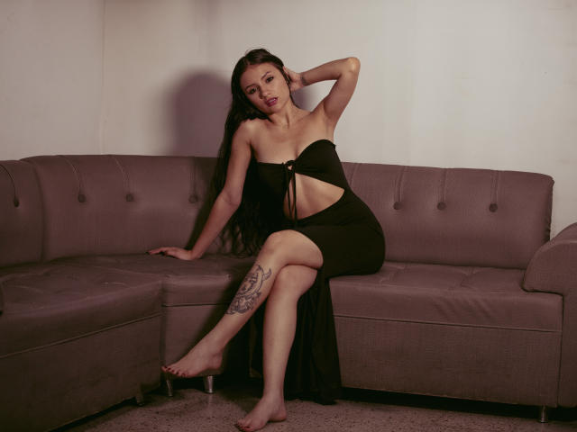 LizzeJoy, with long black hair, wearing a black strapless dress, sits on a gray sofa, her arm behind her head, showing a thigh tattoo, barefoot, looking seductive.