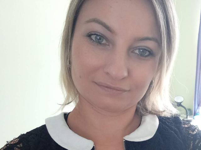 AngelaSamt has blue eyes, light brown hair, and a subtle smile. She's wearing a black lace top with a white collar, showing her tits slightly. She's looking at the camera.