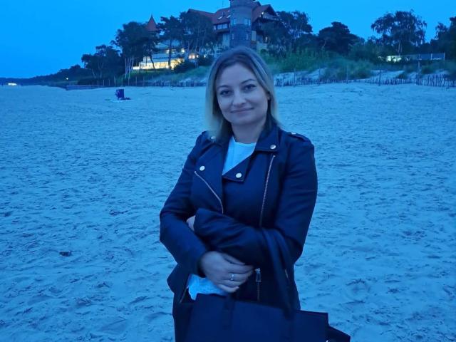 AngelaSamt stands on a beach, arms crossed, wearing a black leather jacket and white shirt, with blue-tinted skin and blonde hair, smiling slightly.
