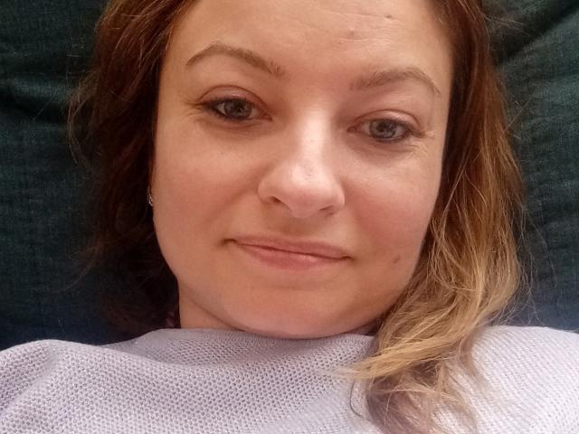AngelaSamt's face with light brown hair, subtle smile, and small nose, wearing a white textured top, lying on a dark green surface. Her eyes are slightly upturned.
