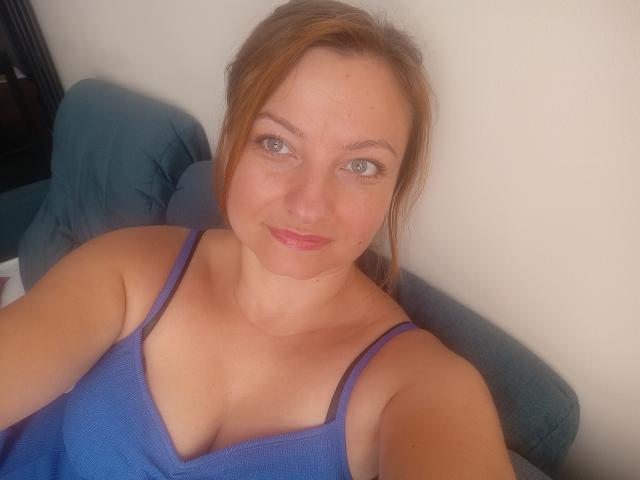 AngelaSamt with blue tank top, showing tits, smiling, light skin, green eyes, red lipstick, brown hair in a ponytail, sitting on a blue couch, looking at the camera.