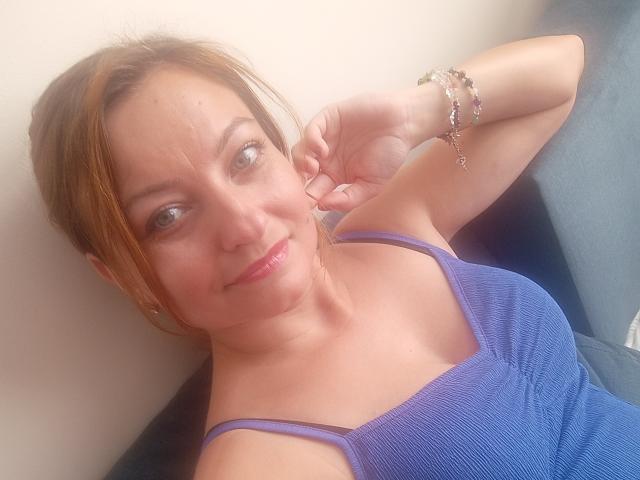 AngelaSamt with light brown hair, wearing a purple ribbed tank top, smiling at the camera, showing small tits, light skin, and a bracelet on her right wrist.