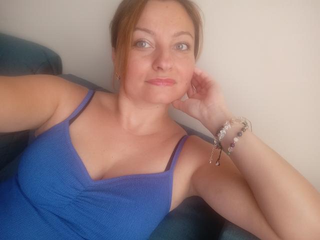 AngelaSamt has small tits, wearing a blue ribbed tank top, with light brown hair, a pink lipstick smile, and a bracelet on her right wrist, sitting on a green chair.