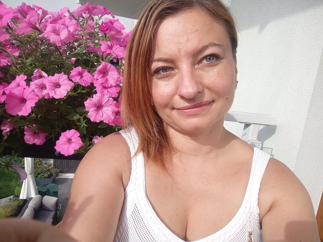 AngelaSamt with light skin, blue eyes, and red-brown hair, wearing a white mesh tank top, showing her boobs, smiling at the camera, pink flowers in the background.