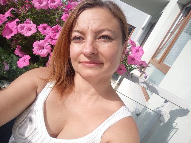 AngelaSamt with light brown hair, white tank top, showing her boobs, pink flowers in background, smiling, fair skin, slight wrinkles, sunlight, medium-sized tits, casual selfie.
