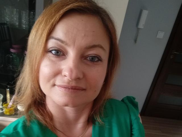 AngelaSamt with light brown hair, blue eyes, and green top, showing slight smile, small nose, and visible wrinkles on forehead, standing in a room with a white phone.