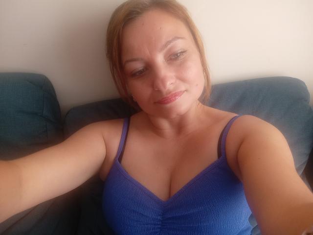 AngelaSamt with light brown hair, blue spaghetti-strap top, showing her tits, slightly smiling, sitting on a dark blue couch, holding a phone for a selfie.