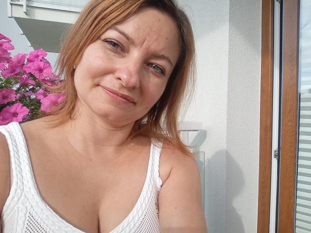 AngelaSamt with light brown hair, white mesh tank top showing her big boobs, slight smile, light skin, and pink flowers in the background, standing by a window.