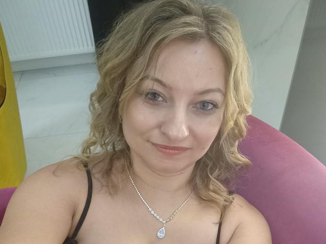 AngelaSamt has curly blonde hair, blue eyes, and is smiling slightly with pink lips. She's wearing a black spaghetti strap top and a silver necklace with a pendant.