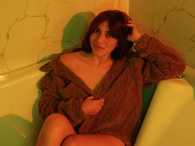 SpencerLe with red hair, sitting in a bathtub, wearing a brown, fuzzy off-shoulder sweater, exposing her tits, looking at the camera with a slight smile.