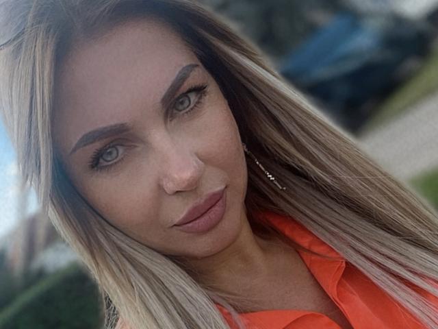 AmandaSexyX, con capelli biondi, occhi verdi e un piercing al naso, indossa una maglietta arancione, ha labbra piene e un leggero sorriso, e guarda direttamente la fotocamera..