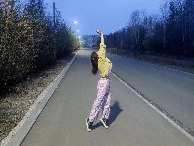 Barbariana with brown hair, yellow jacket, and pink floral pants, standing on a road, arms raised, looking back, wearing black and white sneakers, at dusk.