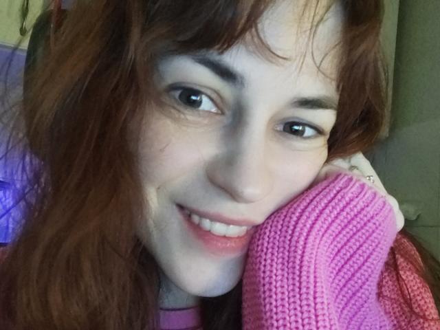Barbariana with red hair, smiling, holding a pink knitted sweater to her face, has light skin and brown eyes, wearing a pink top, looking cute and sexy.