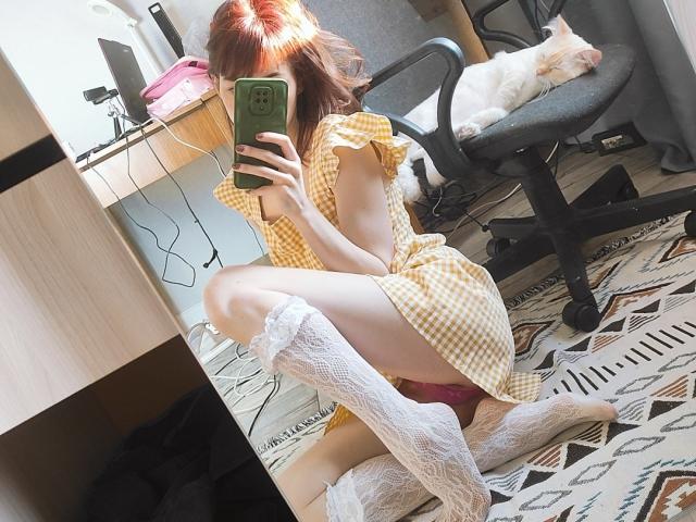Barbariana with red hair, squatting, wearing yellow checkered dress, white lace thigh-highs, taking selfie, holding green phone, chair with sleeping cat, wooden desk, patterned rug, sunlight.