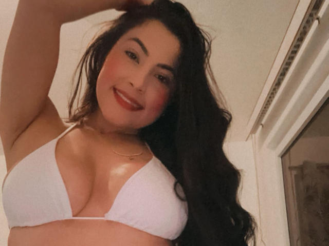 Cryztalx with long black hair, smiling, wearing a white bikini top, showing her big tits, arm raised, red lipstick, and a gold necklace, in a slightly blurry photo.
