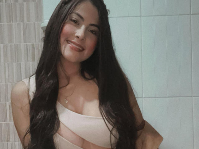 Cryztalx has long dark hair, a smiling face, and is wearing a pinkish-white top that shows her tits. She's standing in a bathroom with white tiles.