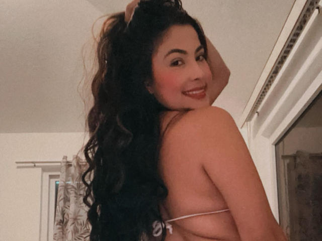 Cryztalx with long black curly hair, smiling at the camera, looking over her shoulder, wearing a white bikini, showing her back and a bit of her tits.