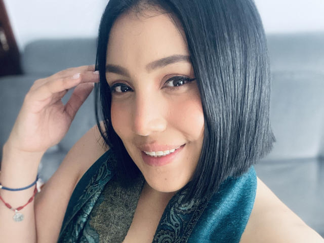 SamantaMuller with black bob, smiling, wearing green patterned scarf, touching hair, brown eyes, subtle makeup, light skin, red bracelet, gray background, soft light, attractive, cute.