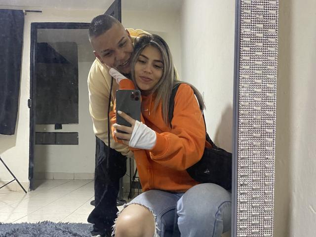 TaylorftDavid in an orange hoodie and jeans, sitting, holding phone, with a man in a yellow shirt and black pants, leaning behind her, taking a mirror selfie.
