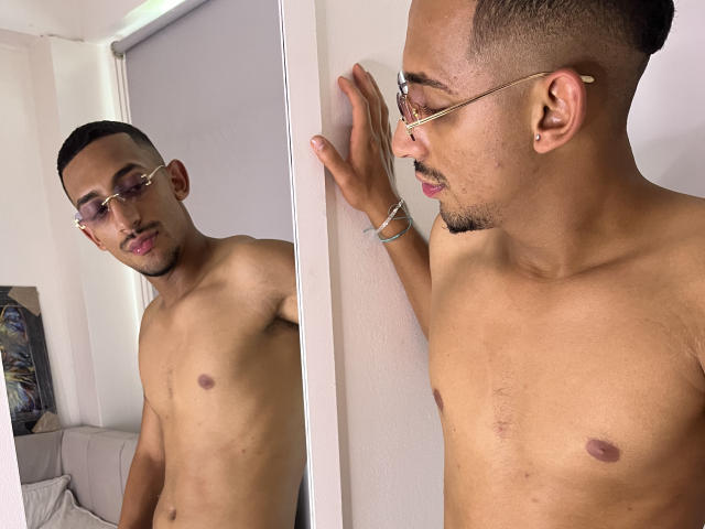 LuckyStar69, torse nu, portant des lunettes, avec des cheveux courts et foncés et une moustache, regardant son reflet dans un miroir. Il porte un bracelet et a une expression faciale subtile..