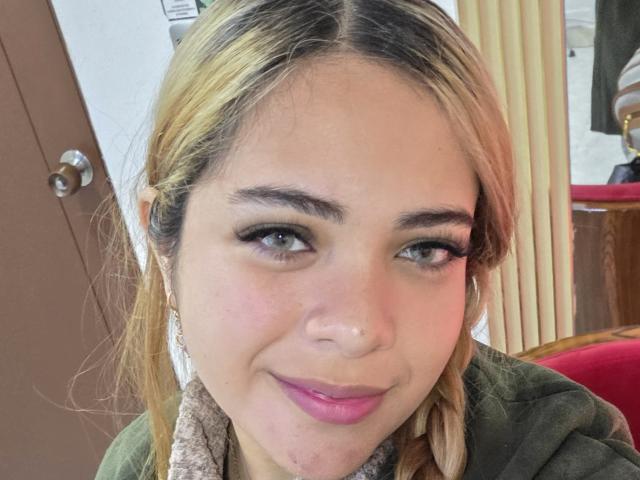 AlicePrice has blonde hair, green eyes, and pink lipstick. She's smiling with a nose piercing, wearing a green jacket and a scarf. Her tits are not visible.