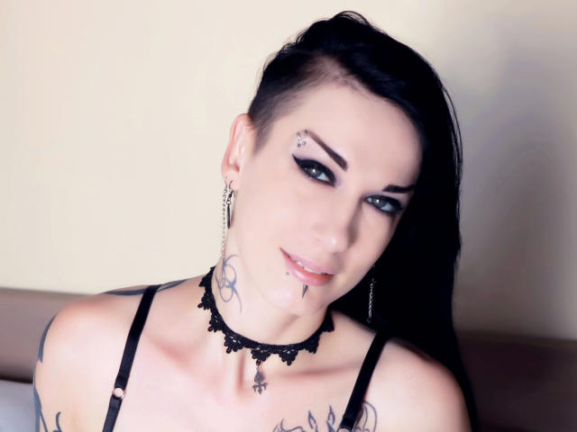 Evelinka-hot ha i capelli neri, tatuaggi e piercing, indossa un collare di pizzo e bretelle nere, ha gli occhi azzurri e un leggero sorriso, e guarda verso la fotocamera..