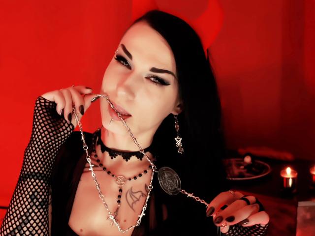 Evelinka, attraente, con maniche a rete nere, occhi rossi, che tiene una catena con un pentagramma, collana nera, corna da diavolo, tatuaggio sul petto, che morde la catena, bagliore rosso, unghie nere, sguardo seducente..