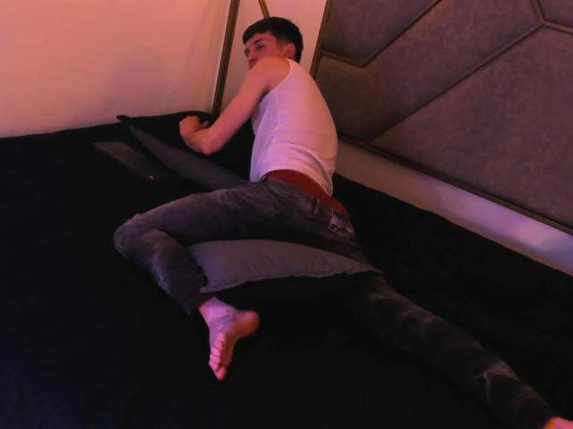 JheamPiero, butt up, wearing a white tank top and gray ripped jeans, lying on a black bed, barefoot, with a red belt, in a dimly lit room.