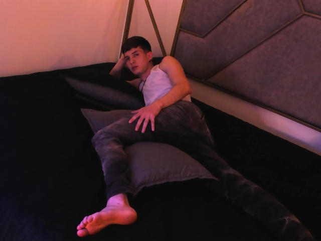 JheamPiero is lying on a black bed, wearing a white sleeveless shirt and gray pants, with his legs spread and bare feet visible, in a dimly lit room.