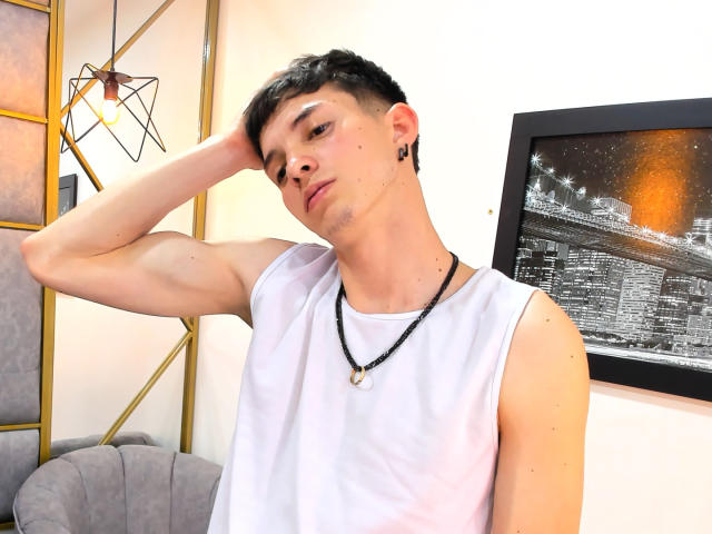 JheamPiero with short black hair, sleeveless white shirt, black necklace, and small ear piercings, hand in hair, looking slightly down, in a brightly lit room.