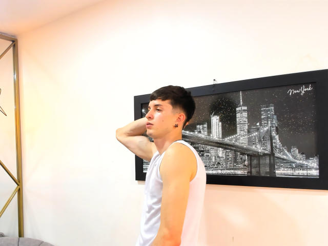 JheamPiero, with short dark hair, wears a white tank top, has a black ear piercing, and stands with arm behind head, looking side, in front of black-and-white city photo.