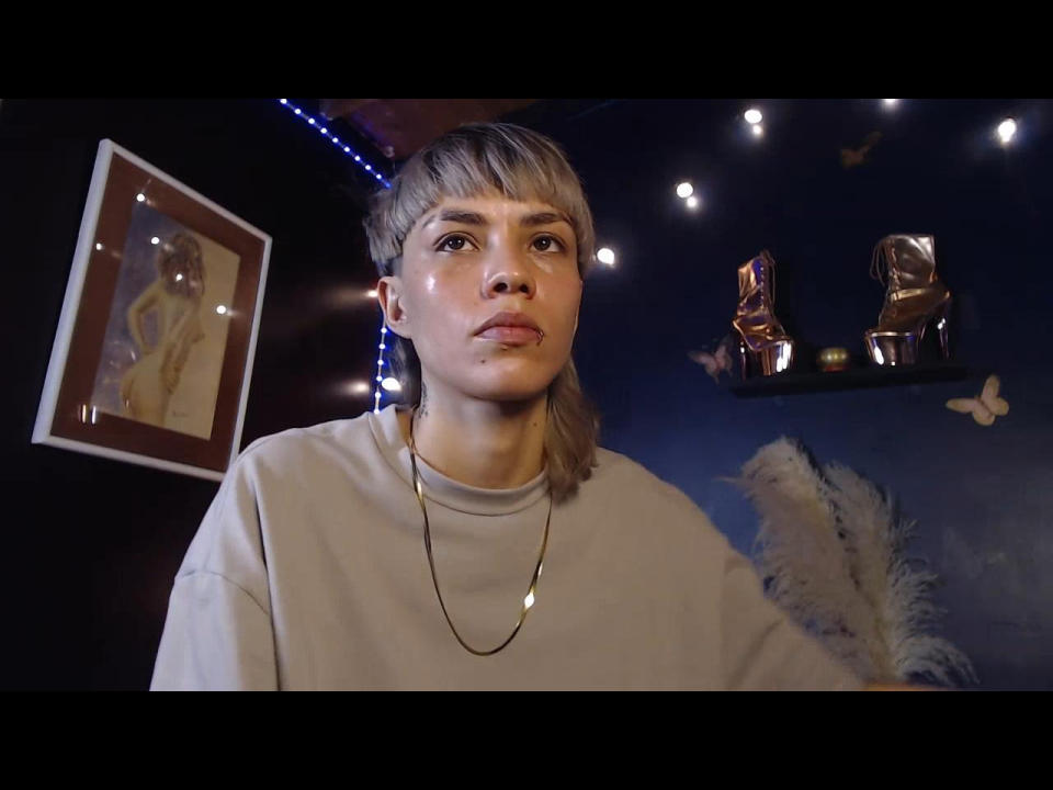 PaytonStorm with short blonde hair, wearing a white sweater and gold necklace, has shiny skin and a serious expression, standing in a dimly lit room with art and sculptures.