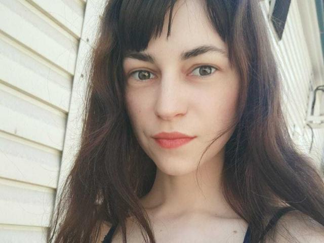 Barbariana has long brown hair, green eyes, and red lips, with a small nose and light skin. She's wearing a black top, looking straight at the camera.