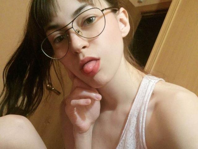 Barbariana with glasses, sticking out her tongue, touching her cheek with her right hand, wearing a white tank top, brown hair, fair skin, in a selfie.