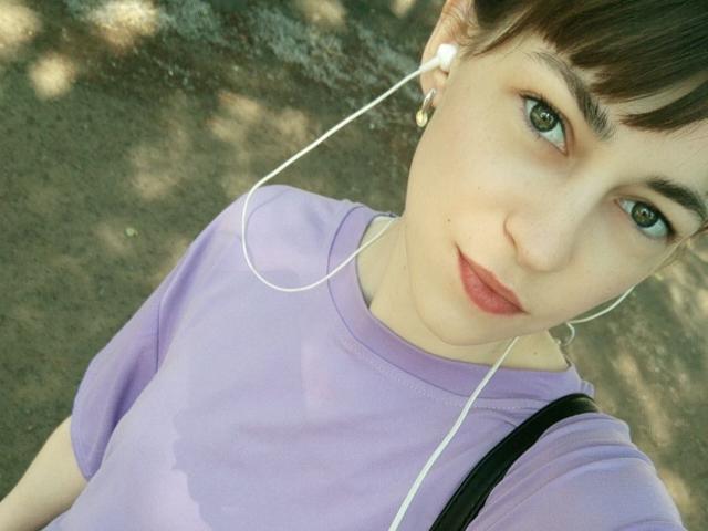 Barbariana has short brown hair, green eyes, and wears a purple shirt with white earbuds. Her small tits are slightly visible. She's looking up at the camera.