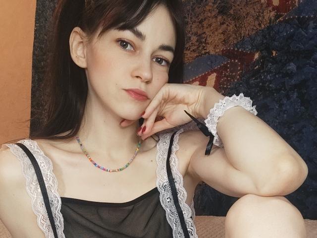 Barbariana with pale skin, dark brown hair in pigtails, wearing a black lace-trimmed top, colorful bead necklace, white lace bracelet, red nails, resting chin on hand, looking at camera.