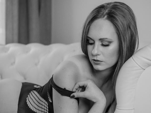 Black-and-white photo of LyraFantasy with long hair, looking down, pulling down a fishnet top, exposing her shoulder and upper arm, resting on a tufted white couch.