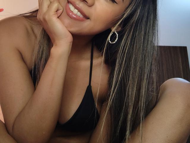 EimyRousse, com cabelos castanhos longos e lisos, vestindo um top de biquíni preto, sorrindo com lábios brilhantes, com a mão no rosto, usando brincos grandes e com os seios levemente visíveis..
