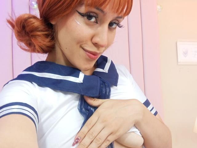 KaylinKrof with orange hair, wearing a blue and white sailor outfit, has her hand over her tits, smiling with winged eyeliner, and a small white face mark.