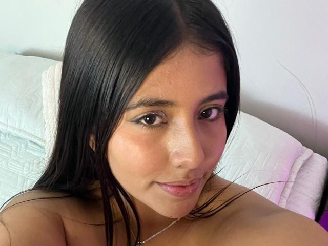 Miiacolliins has long black hair, brown eyes, and a slight smile, showing bare shoulders, with a small silver necklace and white pillow in the background.