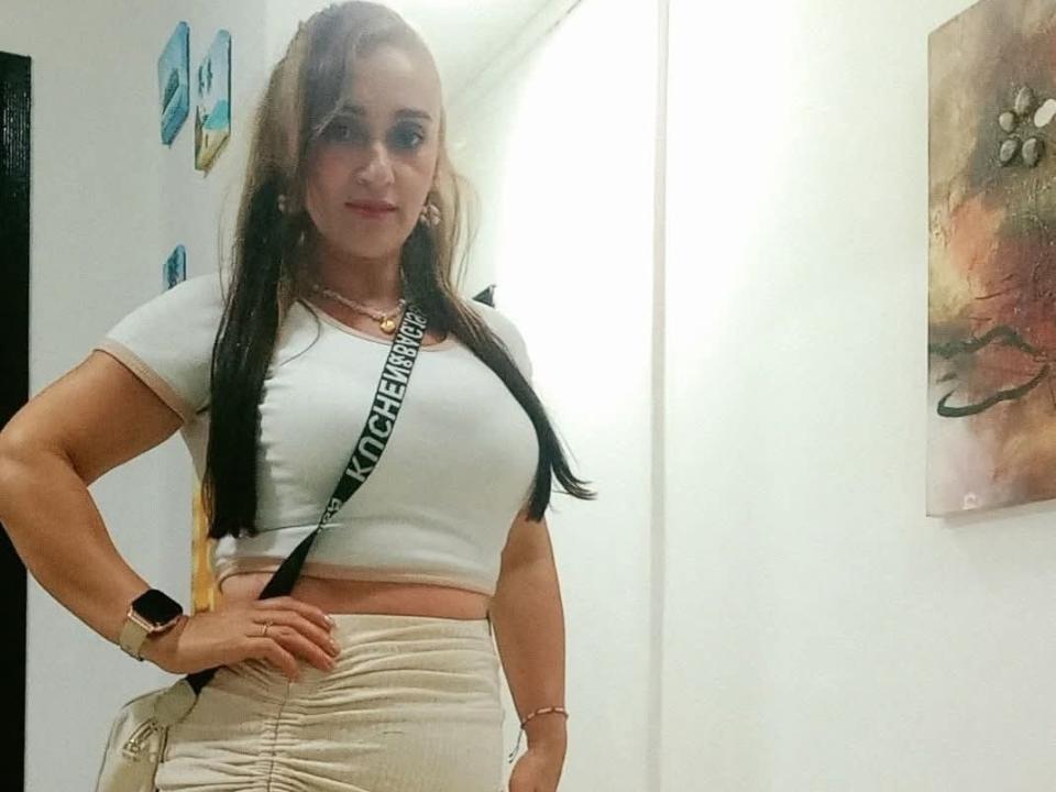 MonaVallen with long brown hair, white crop top, showing her tits, beige skirt, black strap, wristwatch, and pink bracelet, standing in a brightly lit room.