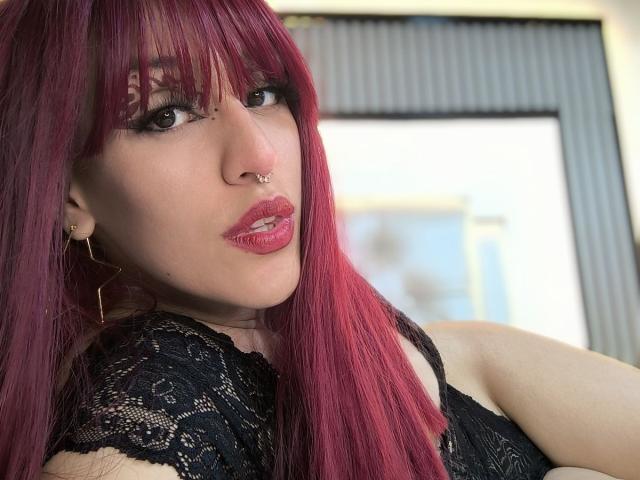 Kaylin Krof, com cabelo ruivo, piercing no nariz e no lábio, vestindo renda preta, tem lábios rosados brilhantes e brincos de argola dourados, olhando para a câmera com um leve franzido..