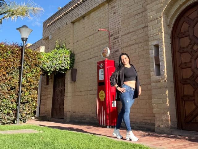 ChristyCuteX stands in front of a red vintage gas pump, wearing a black crop top, blue jeans, and white sneakers, with a black jacket and long hair.