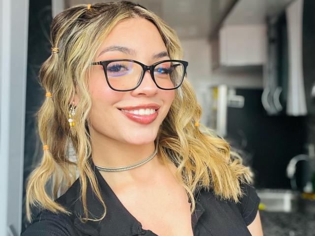 IvyCors with wavy blonde hair, black glasses, and orange hairpins, smiling with pink lipstick, wearing a black shirt and silver choker, showing her teeth and small nose.