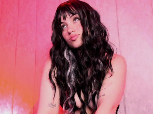 SophieRouge with long, wavy black hair, pinkish-red background, tattoos on arms, pouty lips, looking slightly to the side, topless, showcasing her bare tits.
